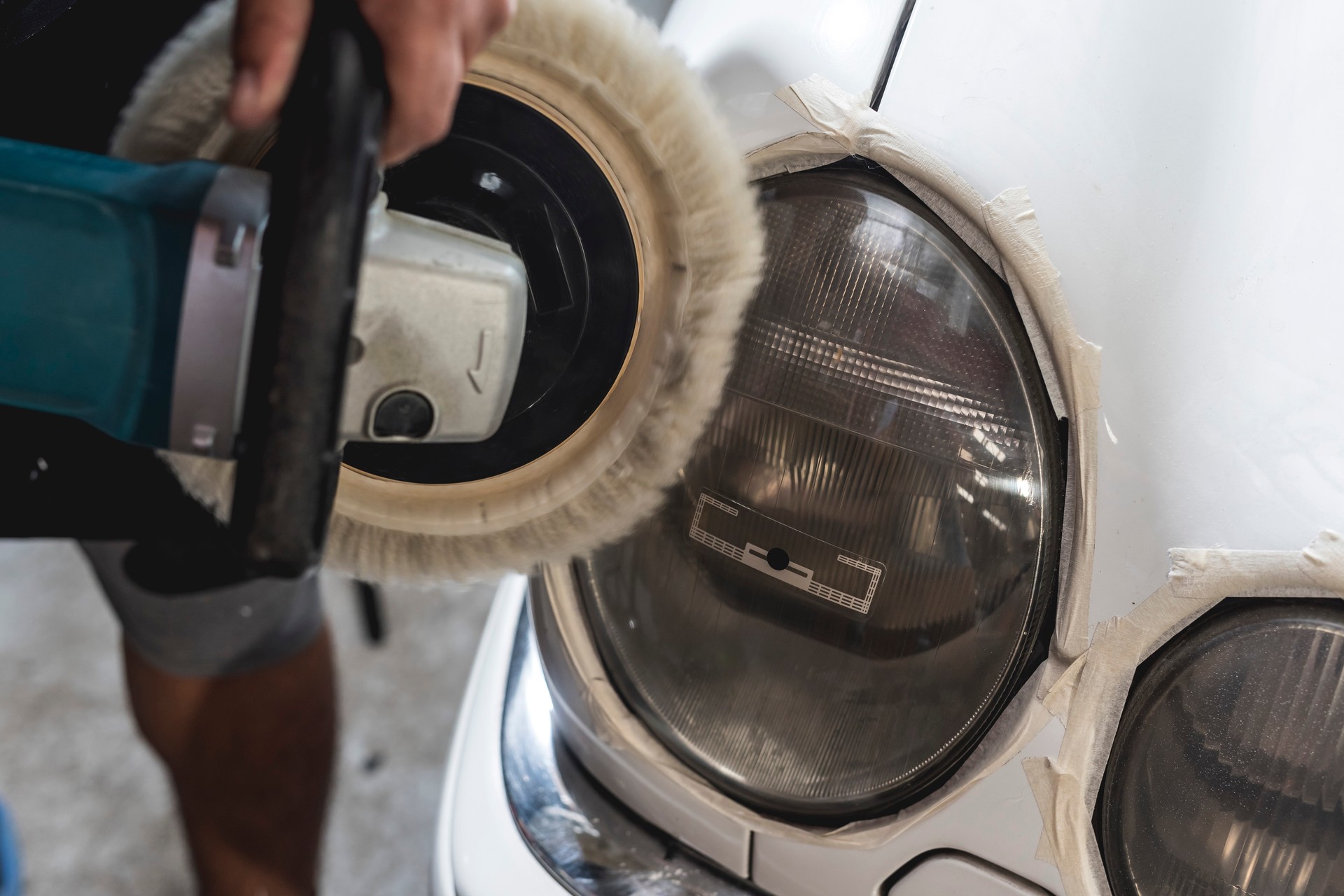 A man buffing the circular headlights of a sedan with a wool pad attached to a rotary. masking tape placed on edges for protection. Restoring foggy headlights at a car repair shop.